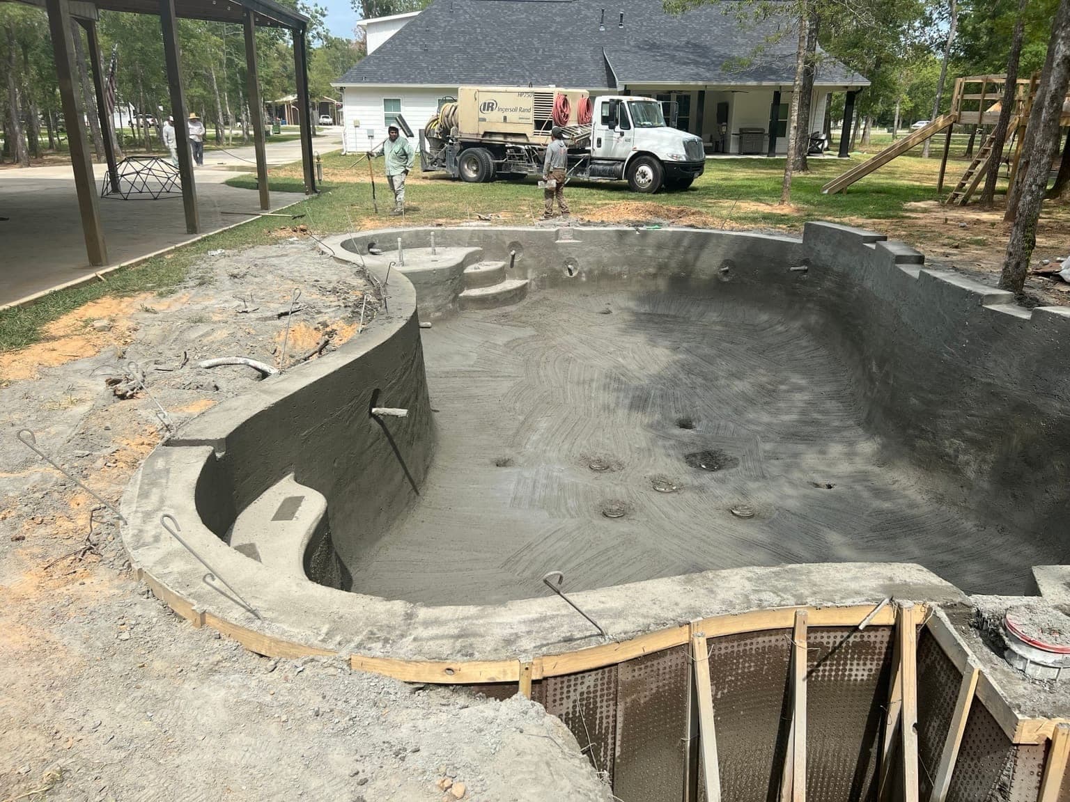 Freeform pool shell just after gunite application with gunite truck and crew visible in background