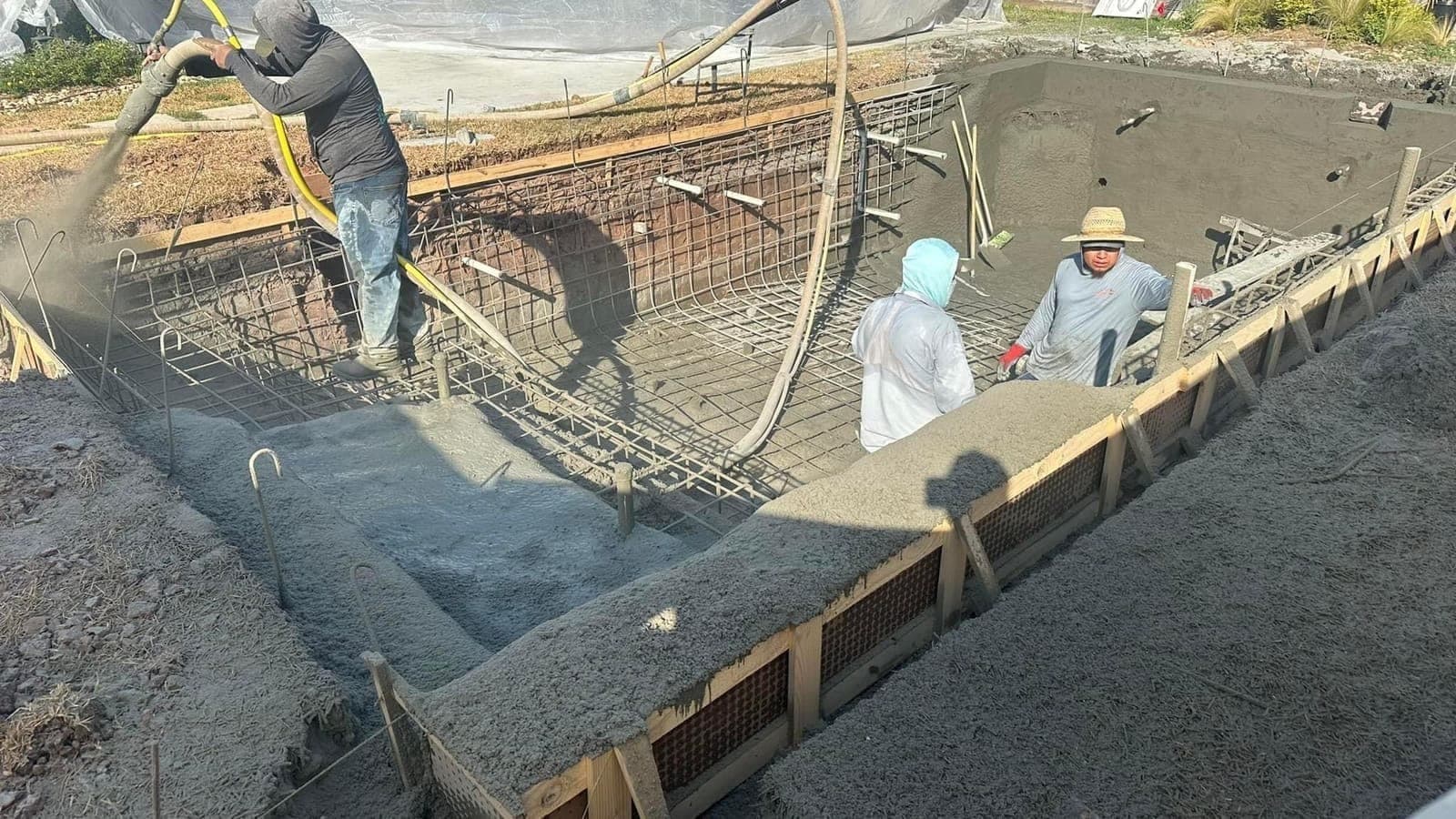 Crew actively shooting gunite on a pool shell, hose arcing overhead, wooden formwork visible