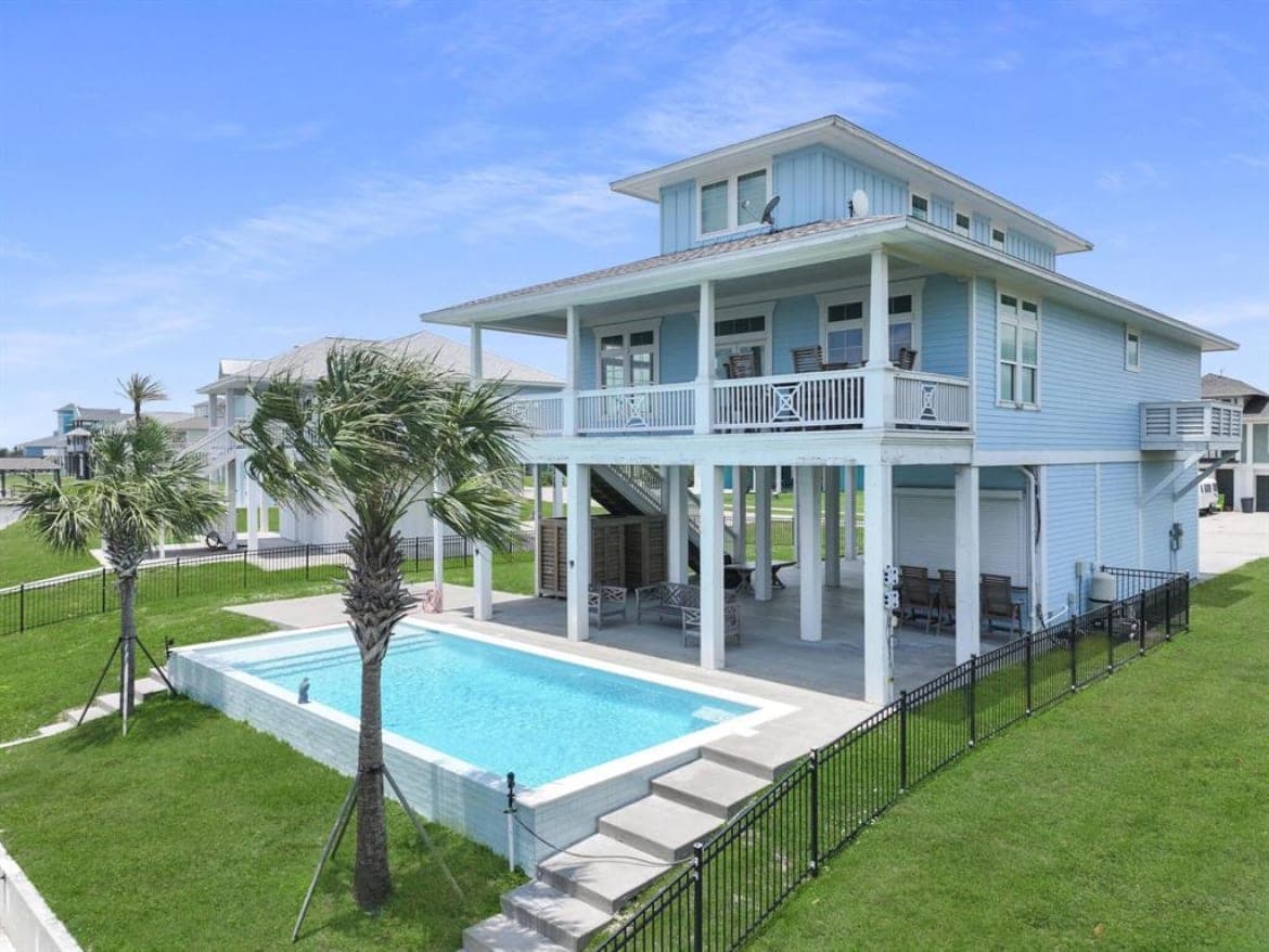 Completed rectangular gunite pool with a tanning ledge beneath an elevated blue Gulf Coast home, framed by palm trees and a fenced lawn